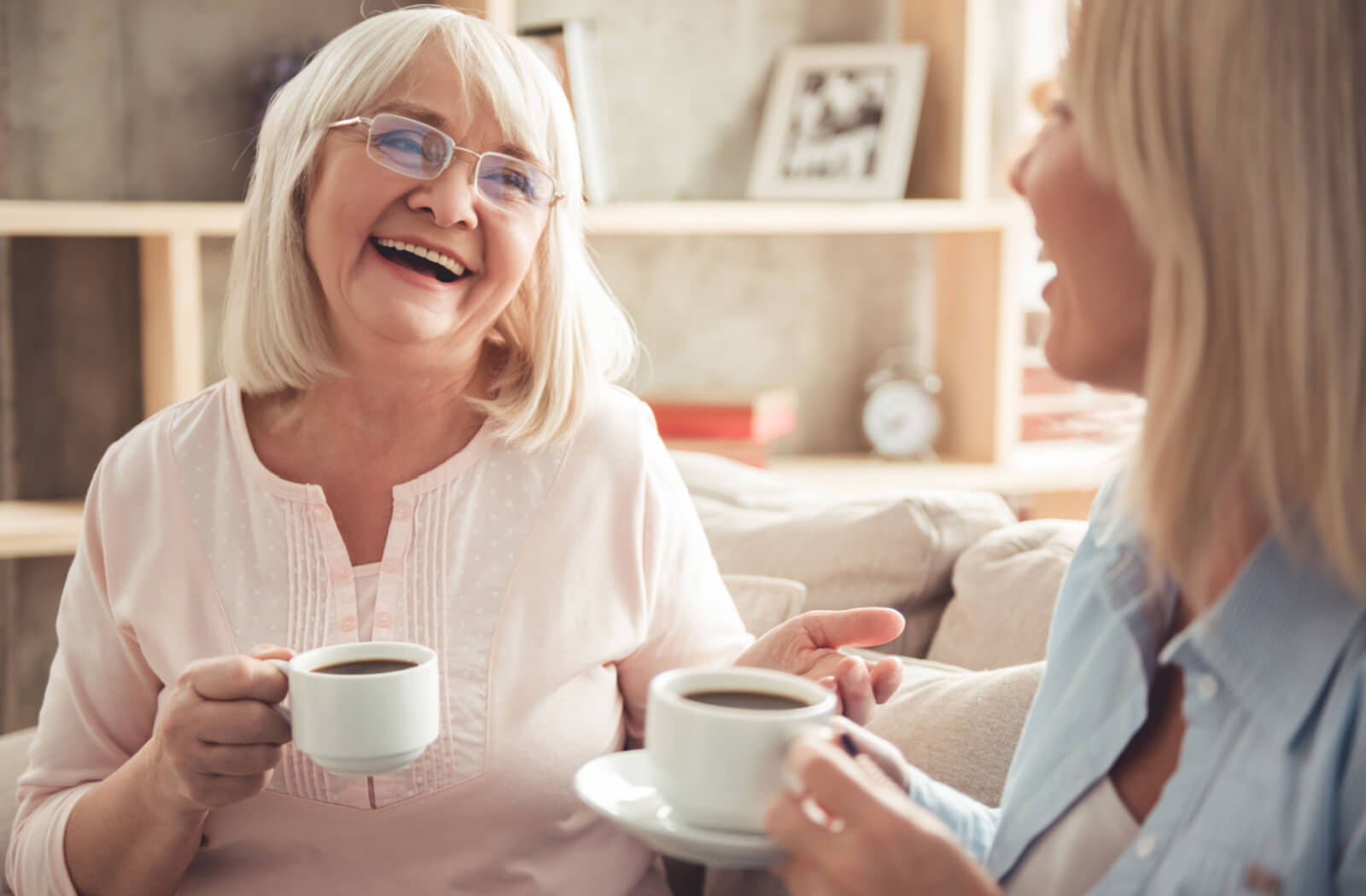 A mother and her daughter sitting on a couch smiling and talking to each other while holding a cup of coffee.
