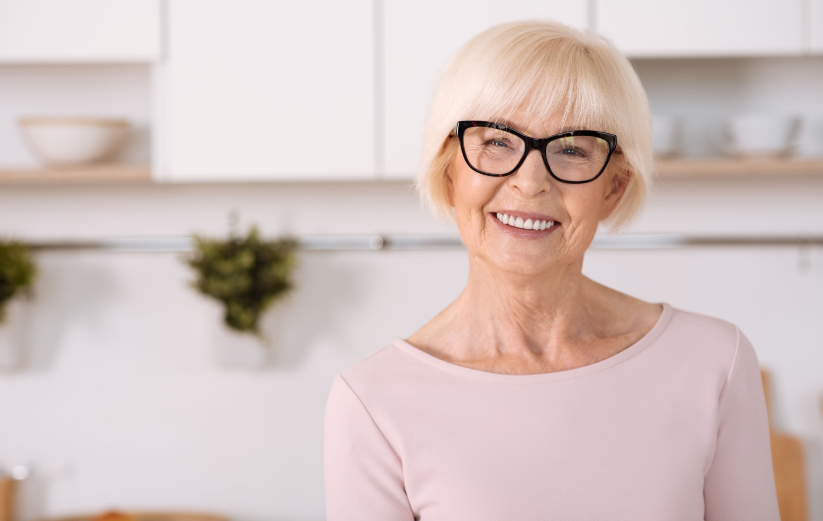 An older adult wearing black, thick-framed glasses smiles at the camera with bright, straight teeth.