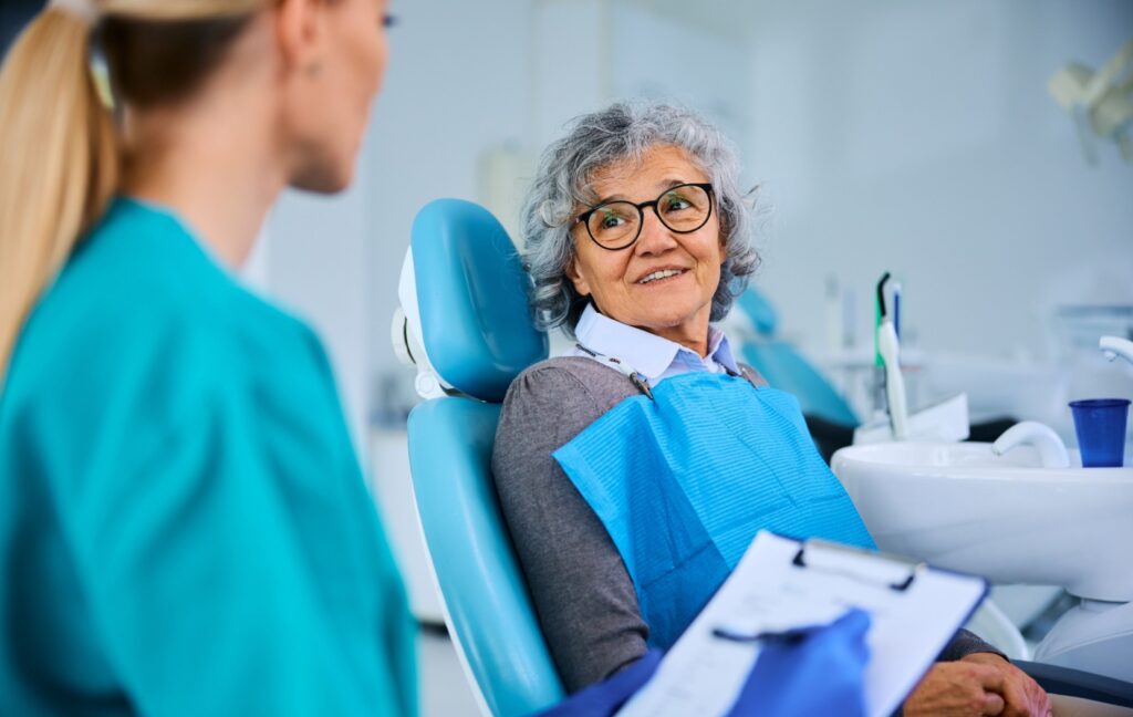 An older adult sits in an examination chair at their dentist's office during a dental exam while their dentist discusses their oral health with them.