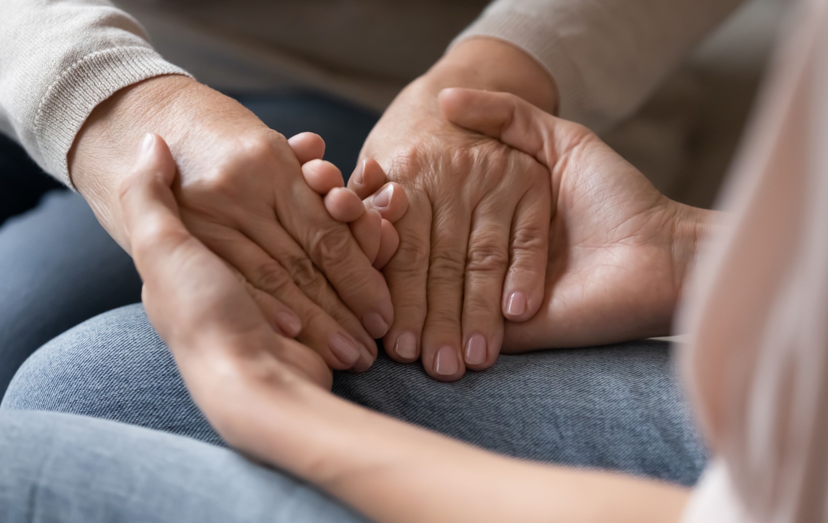 An adult child reaches out both of their hands to hold their elderly parent's hands in their lap.