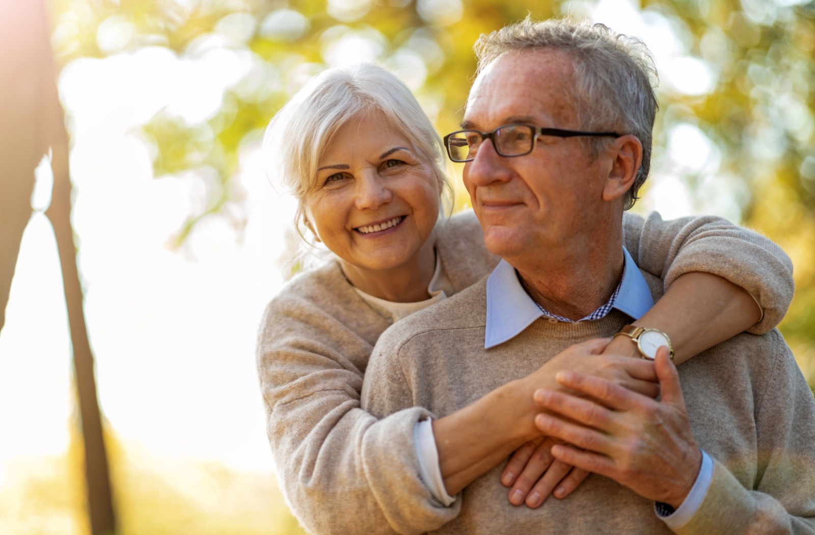 A senior couple embracing each other in a park with the sun shining in the background.